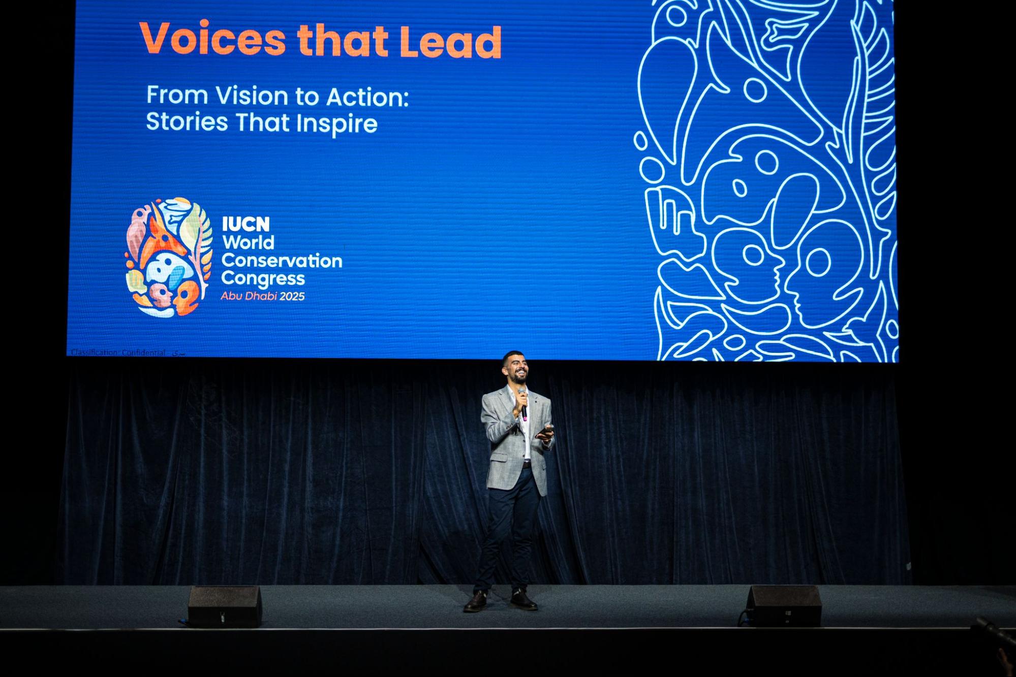 A well-dressed young man speaks on stage, delivering a talk called "Voices that lead."